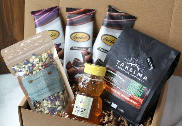 Display of items that can make up a curated gift box, including three bars of Bedré chocolate, one bag of Sakari Farms tea, one bag of Takelma Roasting Co ground coffee and one adorable bear-shaped honey bottle.