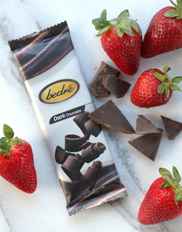 Three wrapped chocolate bars (milk, dark and milk with almonds) are laying flat surrounded by strawberries on a marble background.