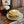 Load image into Gallery viewer, A stack of delicious looking pancakes with some melting butter on top. They are sitting on a plate on a wood table next to a maple shaped bottle of maple syrup and a bag of the wild rice pancake mix that they are made out of.
