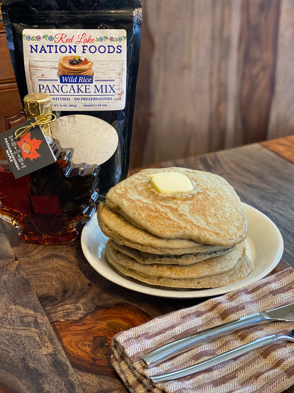 A stack of delicious looking pancakes with some melting butter on top. They are sitting on a plate on a wood table next to a maple shaped bottle of maple syrup and a bag of the wild rice pancake mix that they are made out of.