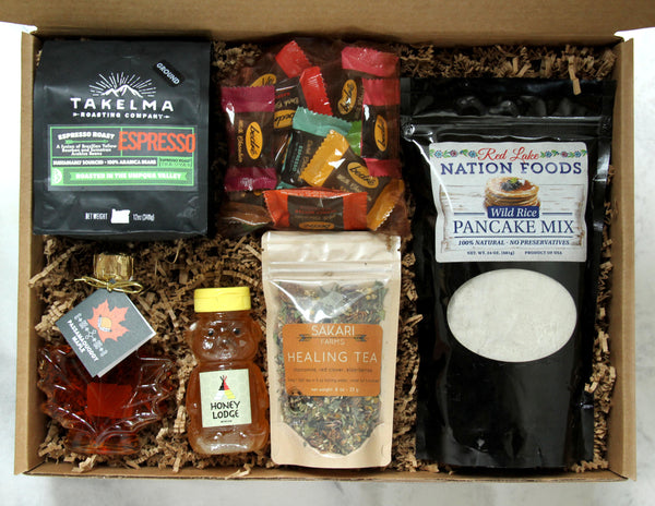 Display of items that can make up a Rise and Resist Box, including one bag of 12 Bedré meltaways, one bag of Sakari Farms tea, one bag of Takelma Roasting Co ground coffee, a maple leaf shaped bottle of maple syrup, a bag of wild rice pancake mix and one adorable bear-shaped honey bottle.