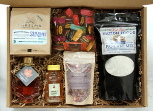 Display of items that can make up a Rise and Resist Box, including one bag of 12 Bedré meltaways, one bag of Sakari Farms tea, one bag of Takelma Roasting Co ground coffee, a maple leaf shaped bottle of maple syrup, a bag of wild rice pancake mix and one adorable bear-shaped honey bottle.