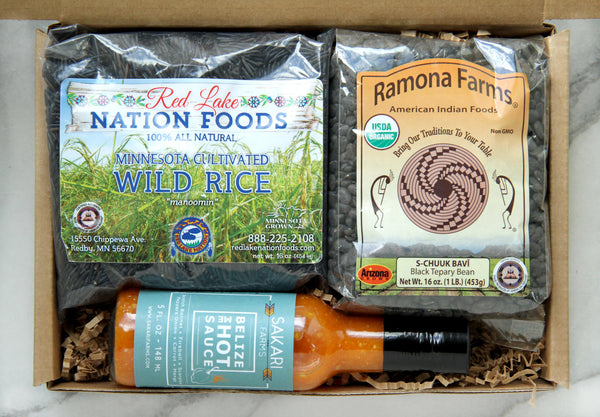Display of items that can make up a Some Like it Hot Box, including one bag of black Ramona Farms Teppary Beans, one bottle of Belize Me hot sauce from Sakari Farms and a bag of Red Lake Nation wild rice.