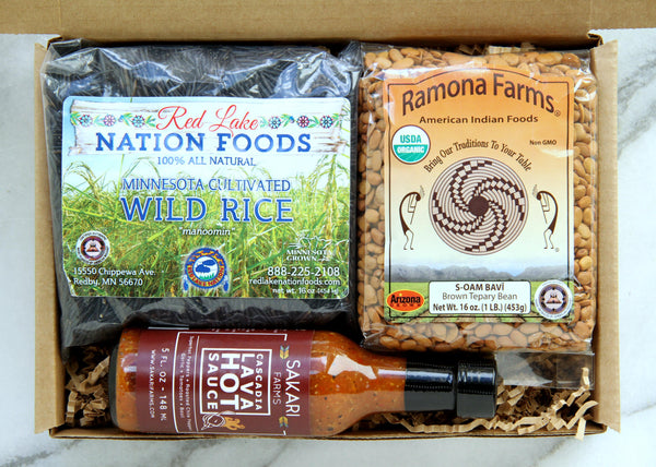 Display of items that can make up a Some Like it Hot Box, including one bag of brown Ramona Farms Teppary Beans, one bottle of Cascadia Lava hot sauce from Sakari Farms and a bag of Red Lake Nation wild rice.