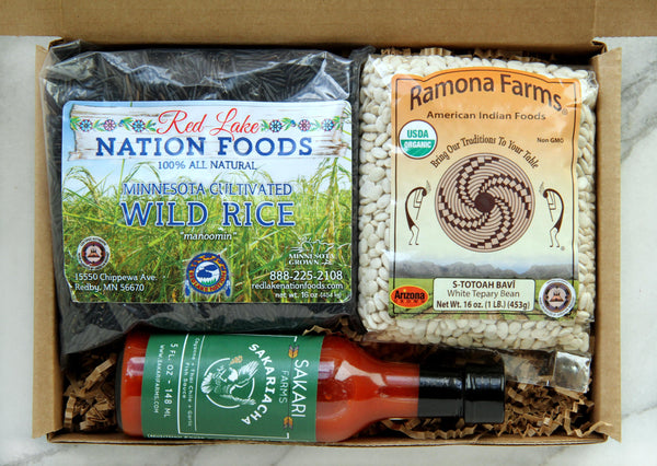Display of items that can make up a Some Like it Hot Box, including one bag of Ramona Farms white Teppary Beans, one bottle of Sakariacha hot sauce from Sakari Farms and a bag of Red Lake Nation wild rice.